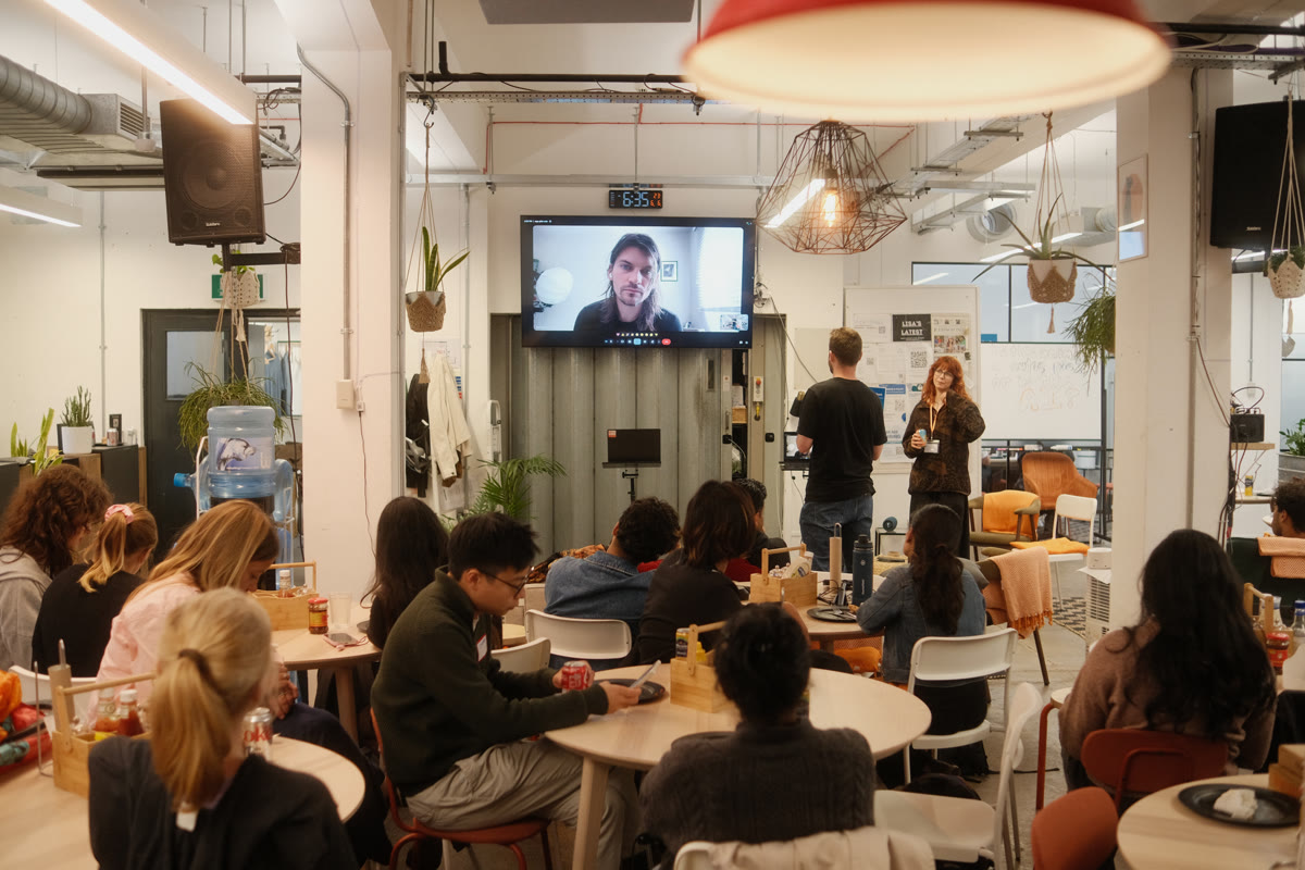 Participants seated at tables watching a presentation at the AI Risk Content Hackathon, with a speaker joining on screen